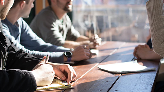 close up angle of three people sitting at a wooden table near a window overlooking the city with pens and notepads; image used for blog post for IRS new regulations for not-for-profits UBIT calculation