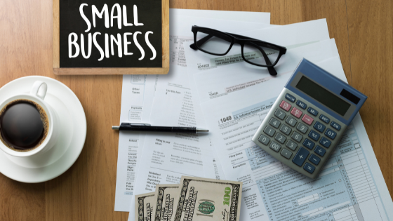 A calculator, pair of reading glasses, money and a chalkboard reading "Small Business," sitting on top of financial documents; image used for blog post about tax breaks for small businesses